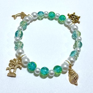 May include: A beaded bracelet featuring white faux pearls and iridescent teal and green beads. Gold-colored charms include a dolphin, a ship's wheel, a palm tree, and a seashell. The bracelet is designed with an ocean or nautical theme.