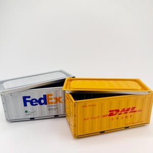 May include: Two miniature shipping container models, one gray with the FedEx logo and the other yellow with the DHL logo. Both containers have removable lids and are set against a white background. The FedEx container has the text "express fedex.com".