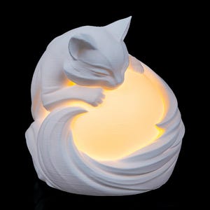 May include: A white cat-shaped lamp with a warm, glowing light emanating from within. The cat is curled up, with its head resting on its paws. The lamp has a textured surface and is set against a black background.