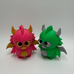 Könnte beinhalten: Zwei kleine Drachenfiguren. Die linke Figur ist leuchtend pink mit goldenen Hörnern, Flügeln und Bauch. Die rechte Figur ist leuchtend grün mit grauen Hörnern, Flügeln und Bauch. Beide haben große schwarze Augen und eine kleine Schnauze mit 3D-Druck-Struktur.