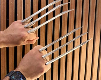 May include: A pair of silver-colored metal claw props, each featuring three long, pointed blades extending from a central handle grip. The blades are straight with beveled edges, designed to be held in the hand for display or costume use.