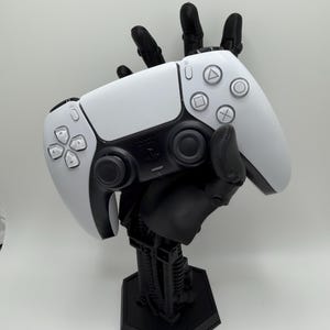 May include: A white and black PlayStation 5 controller held by a black robotic hand-shaped stand. The controller has a symmetrical design with various buttons and a touchpad. The stand has a detailed mechanical design.