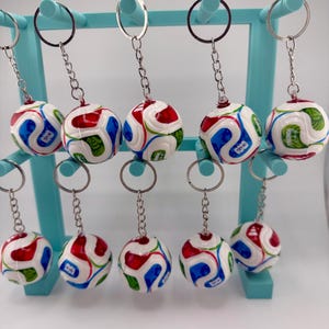 Può includere: Una collezione di portachiavi a forma di pallone da calcio. Ogni portachiavi presenta una piccola sfera con base bianca e disegni curvi rossi, blu e verdi. I portachiavi sono esposti su un supporto azzurro con anelli argentati.