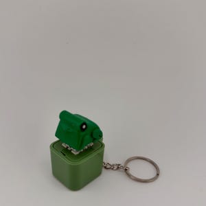 Könnte beinhalten: Ein grüner, dinosaurierförmiger Schlüsselanhänger mit quadratischer Basis. Der Dinosaurier hat ein kleines Auge und Zähne. Der Schlüsselanhänger ist an einem silbernen Metallring befestigt.