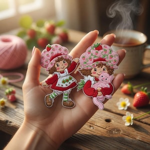 May include: Two embroidered patches featuring a cartoon girl with a strawberry hat and dress. One patch shows the girl with arms raised, the other holding a pink cat. The patches are held in a hand, with strawberries and a cup of tea in the background.