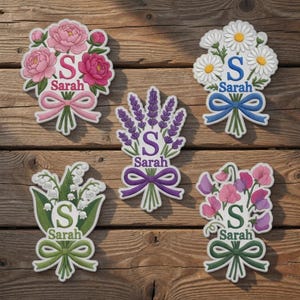 Op de afbeelding: Vijf bloemboeketontwerpen met de naam "Sarah" en de letter "S". De boeketten bevatten pioenrozen, madeliefjes, lavendel, lelietjes-van-dalen en lathyrus. Elk boeket heeft een lint en staat op een houten achtergrond.
