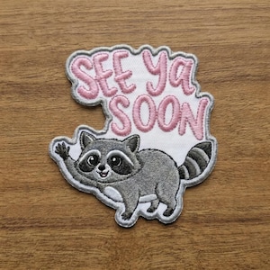 May include: Embroidered patch featuring a gray raccoon waving with the text "SEE YA SOON" in pink. The patch has a white background and a gray border, perfect for adding to clothing or accessories.