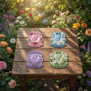 Op de afbeelding: Vier geborduurde patches met bloemmotieven en de letter "S" worden getoond op een houten tafel. De patches zijn in roze, blauw, paars en groen, elk met een ander bloemarrangement en de naam "Sarah".