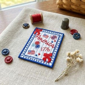 May include: Embroidered patriotic patch with the word "America" and the year "1776" in red script. The patch features American flags, fireworks, and bows. Additional items include a red spool of thread, buttons, and a thimble.