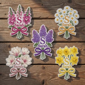 Op de afbeelding: Een verzameling bloemendecoraties, elk met een andere bloem: vingerhoedskruid, madeliefjes, seringen, magnolia's en narcissen. Elk ontwerp bevat de letter "S" en de naam "Sarah". De bloemen zijn vastgebonden met een lint.