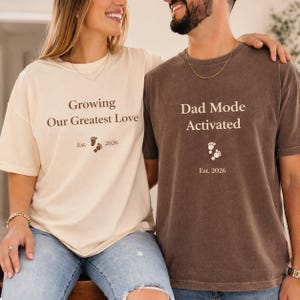 Puede incluir: Dos camisetas a juego, una crema y otra marrón, con texto de anuncio de embarazo. La camiseta crema dice "Growing Our Greatest Love" con huellas de bebé y "Est. 2026". La camiseta marrón dice "Dad Mode Activated" con huellas y "Est. 2026."
