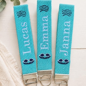 May include: Three turquoise fabric key fobs with silver-toned metal hardware and split rings. Each strap features embroidered dark blue wave icons, light blue names reading Lucas, Emma, and Janna, and dark blue swimming goggles at the bottom.