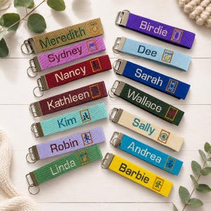 May include: A collection of rectangular fabric keychains in various colors including yellow, purple, blue, green, and red. Each keychain features embroidered names such as Meredith, Sydney, Nancy, Kathleen, Kim, Robin, Linda, Birdie, Dee, Sarah, Wallace, Sally, Andrea, and Barbie.