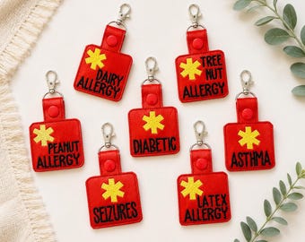 Puede incluir: Siete llaveros médicos cuadrados rojos con cierres de mosquetón plateados y broches a presión. Cada uno presenta un símbolo de Estrella de la Vida bordado en amarillo y texto negro que dice Dairy Allergy, Tree Nut Allergy, Peanut Allergy, Diabetic, Asthma, Seizures o Latex Allergy.
