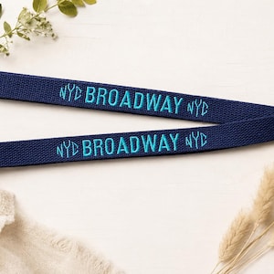 Peut inclure: Un cordon en tissu bleu marine avec le texte brodé BROADWAY en bleu clair, encadré par les lettres NYC de chaque côté. Le cordon est doté d'un mousqueton pivotant en métal argenté à l'extrémité pour attacher des objets.