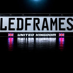 May include: A rectangular black license plate frame featuring the white text LEDFRAMES in a bold font. Below this, the text UNITED KINGDOM is displayed in white, flanked by two small Union Jack flag icons. The frame emits a bright white LED glow.