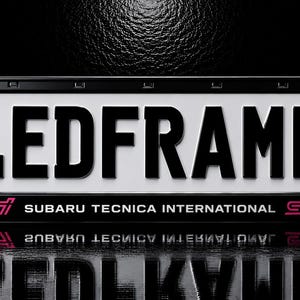 Op de afbeelding: Een zwarte kentekenplaathouder met de tekst LEDFRAMES in grote zwarte letters op een witte achtergrond. Het frame bevat een blauwe Europese Unie-strip met de letter D en het Subaru Tecnica International-logo in roze aan de onderkant.
