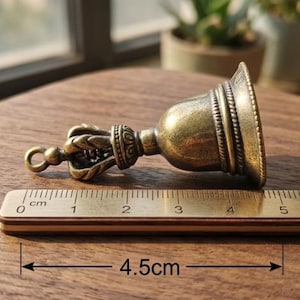 May include: A small, antique-style brass bell, measuring 4.5 cm in length. The bell features intricate detailing on the handle and rim, with a loop for hanging. The bell has a tarnished, aged appearance, suggesting it is vintage or has an antique finish.