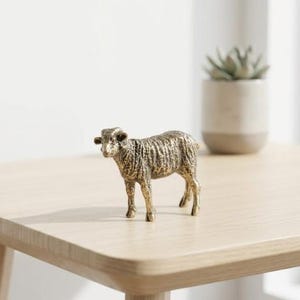 May include: A small, gold-colored sheep figurine with detailed texture. The decorative lamb is standing on a light-colored wooden surface. A potted succulent plant is in the background.