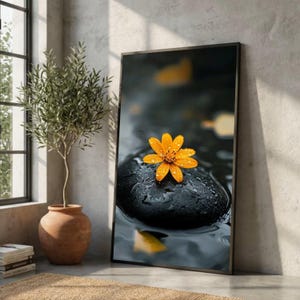 Peut inclure: Une impression d'art mural verticale encadrée représentant une fleur jaune vive avec des gouttelettes d'eau reposant sur une pierre noire lisse et humide. L'arrière-plan est une surface d'eau sombre et floue avec des reflets jaunes doux.