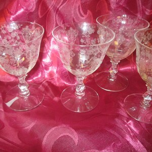 May include: Four clear, stemmed wine glasses with etched floral patterns. The glasses have a rounded bowl, a twisted stem, and a circular base. The glasses are arranged on a pink fabric surface.
