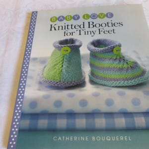 May include: A book titled "Baby Love Knitted Booties for Tiny Feet" by Catherine Bouquerel. The cover features a pair of knitted baby booties in green and blue on a blue and white polka dot fabric.