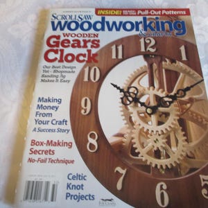 May include: A magazine cover for "Scroll Saw Woodworking & Crafts" featuring a wooden gears clock. The clock has a dark brown wooden frame with white numbers and light-colored wooden gears. The magazine includes articles on woodworking projects.