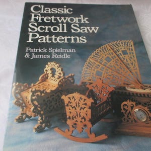 May include: The cover of a book titled "Classic Fretwork Scroll Saw Patterns" by Patrick Spielman & James Reidle. The cover features several examples of fretwork scroll saw projects, including a bed, a cradle, and a fan.