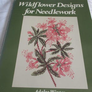May include: A green book titled "Wildflower Designs for Needlework" with a floral cross-stitch design on the cover. The design features pink flowers and green leaves. The author's name, Adalee Winter, is at the bottom.
