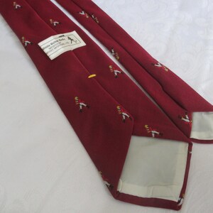May include: Maroon tie with a repeating pattern of figures in red coats and yellow accents. A label with text is visible. The tie is made of a smooth fabric and is suitable for formal occasions.