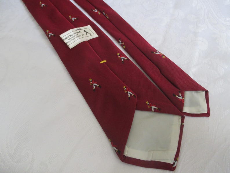 May include: Maroon tie with a repeating pattern of figures in red coats and yellow accents. A label with text is visible. The tie is made of a smooth fabric and is suitable for formal occasions.