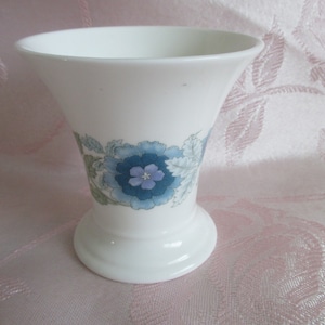 May include: A white ceramic vase with a floral design featuring blue and green flowers. The vase has a flared top and a wide base.