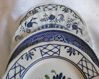 May include: A stack of white ceramic plates with blue floral and geometric patterns. The plates have scalloped edges and varying designs, including stylized flowers and lattice-like borders. The plates are arranged in a slightly overlapping manner.