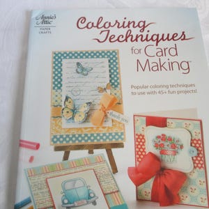 May include: A book titled "Coloring Techniques for Card Making" by Annie's Attic. The cover features examples of colorful handmade cards with butterflies, a car, and floral designs. The book promises over 45 fun projects.