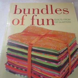 May include: A book titled "bundles of fun" with a stack of colorful fabric fat quarters tied with an orange ribbon. The fabric is a mix of patterns and colors, including orange, green, black, pink, and red. The book cover is light yellow.