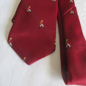 May include: A burgundy tie with a repeating pattern of small figures in yellow hats and red jackets. The tie is made of a textured fabric and is displayed on a white surface. A classic accessory.