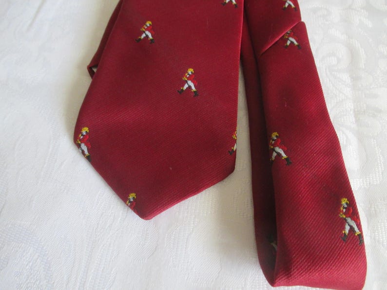 May include: A burgundy tie with a repeating pattern of small figures in yellow hats and red jackets. The tie is made of a textured fabric and is displayed on a white surface. A classic accessory.