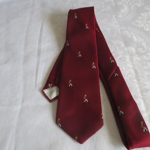 May include: A burgundy tie with a repeating pattern of small figures in red coats and white pants. The tie is made of a smooth fabric and has a classic shape. The background is a white, patterned surface.