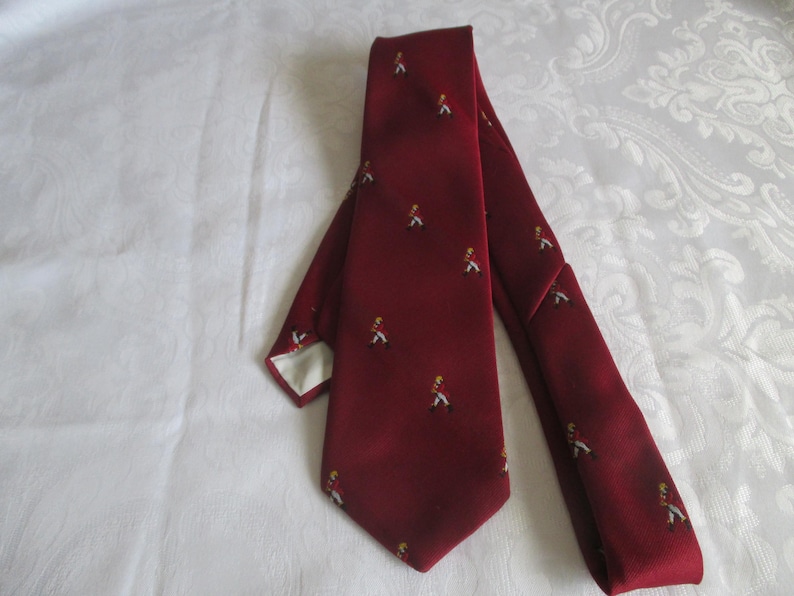 May include: A burgundy tie with a repeating pattern of small figures in red coats and white pants. The tie is made of a smooth fabric and has a classic shape. The background is a white, patterned surface.