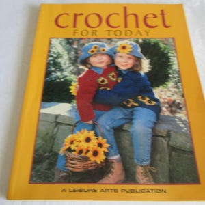 May include: A yellow book titled "crochet FOR TODAY" features two children wearing blue hats and sweaters with sunflower designs. They are holding a basket of sunflowers. A Leisure Arts Publication.
