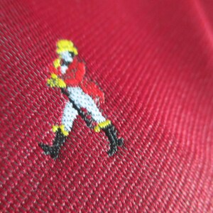 May include: Close-up of a red fabric with a small embroidered figure. The figure is dressed in a red coat, white pants, black boots, and a yellow hat. The fabric has a textured appearance.