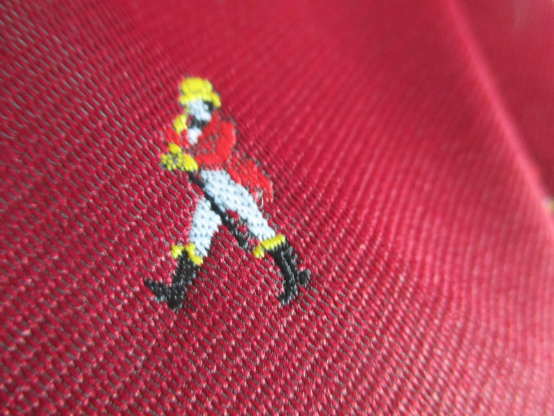 May include: Close-up of a red fabric with a small embroidered figure. The figure is dressed in a red coat, white pants, black boots, and a yellow hat. The fabric has a textured appearance.