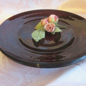 May include: A dark red glass plate with a decorative swirl pattern. A small bouquet of pink fabric roses with green leaves is placed on the plate.