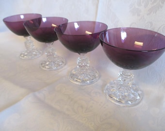 May include: Four purple glass champagne coupes with clear crystal stems and bases. The glasses are arranged in a row on a white tablecloth.