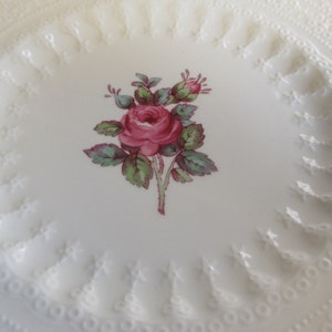 May include: White ceramic plate with a raised, embossed border. The center of the plate features a pink rose with green leaves.