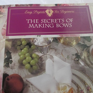 May include: A book titled "The Secrets of Making Bows" with a purple cover and gold lettering. The image includes a table setting with grapes, a peach, and a teacup. The background has a floral pattern.