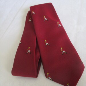 May include: A burgundy tie with a repeating pattern of small figures in red coats and yellow hats. The tie is folded and lies on a white surface. The tie is a classic accessory.