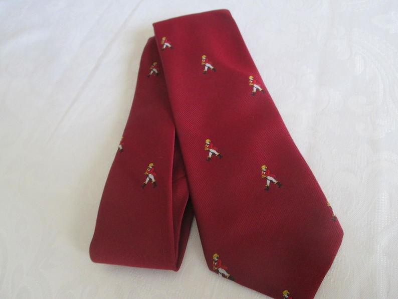 May include: A burgundy tie with a repeating pattern of small figures in red coats and yellow hats. The tie is folded and lies on a white surface. The tie is a classic accessory.