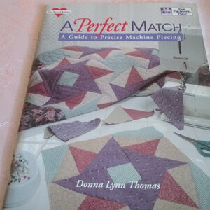 May include: A book titled "A Perfect Match: A Guide to Precise Machine Piecing" by Donna Lynn Thomas. The cover features a quilt with a star pattern in shades of pink, purple, and green. The book is about quilting and sewing.