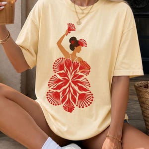 May include: A pale yellow short-sleeve t-shirt featuring a graphic print of a figure with a dark bun hairstyle, holding a red fan, and wearing a large, stylized skirt composed of multiple red floral-like shapes. The shirt has a crew neckline.